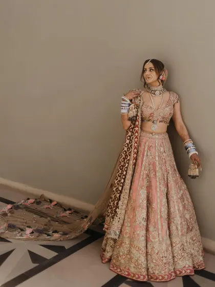 An elegant full-length shot showcasing her unique bridal style. The soft, diffused lighting highlights the delicate embroidery of her lehenga and the flawless finish of her makeup.