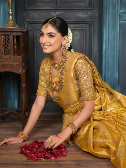 A full-length shot of the bride seated gracefully. The golden saree, temple jewellery, and radiant makeup come together to create a look of pure, timeless elegance.