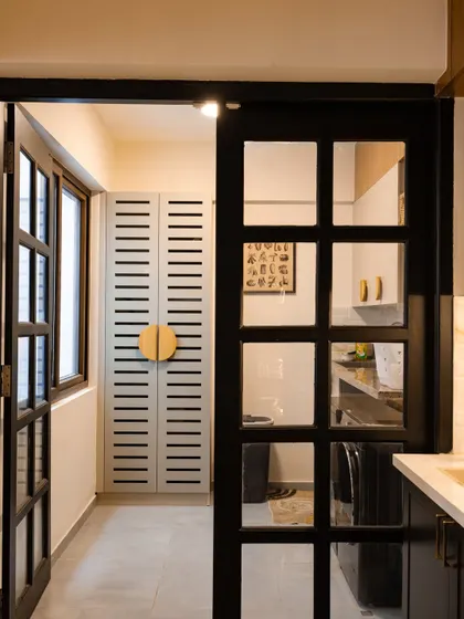 A view from the kitchen into the utility area, separated by a stylish black-framed glass door. The louvered utility cabinet with a large circular handle is a unique design feature.