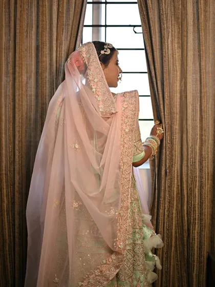A classic bridal pose, looking out the window. The soft light highlights the dewy finish of her skin and the intricate details of her pastel outfit.