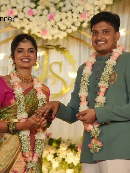A smiling couple at their engagement ceremony, with a backdrop of white flowers, golden arches, and hanging bird motifs.