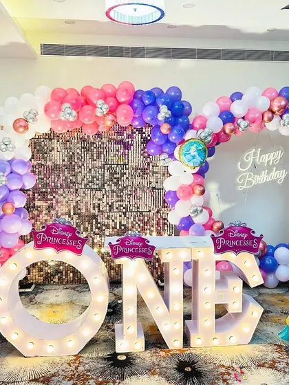 A Disney Princess first birthday party with "ONE" marquee letters. The stage includes a pink sequin wall and cutouts of Aurora, Belle, and Ariel.
