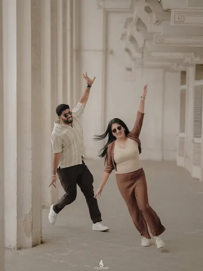 A fun, high-energy pose in a corridor with classic architectural columns. Their coordinated brown and beige outfits are stylish and modern.