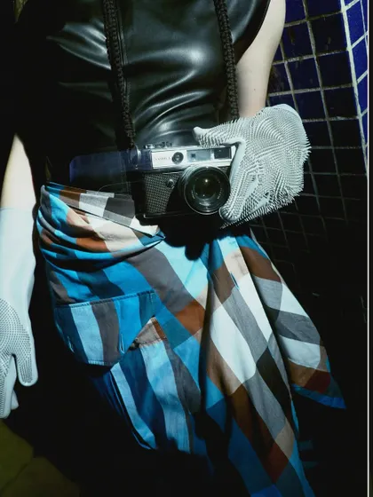 A close-up detail shot from the 'Peek-a-boo' series, focusing on the model holding a vintage camera. The image plays with the idea of the photographer becoming the subject, wrapped in a moody, noir aesthetic.