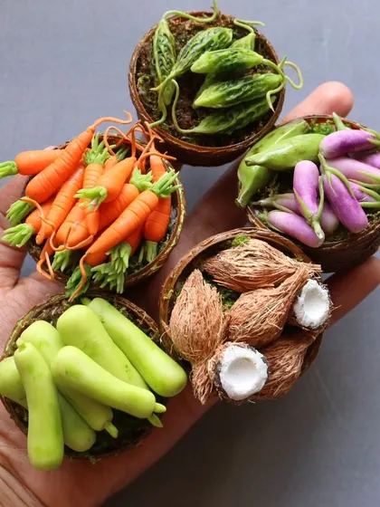 A hand holding five baskets of miniature vegetables, including carrots, brinjals, and coconuts.