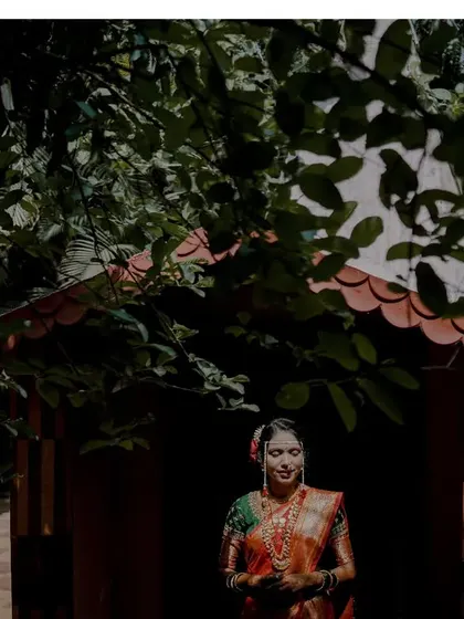 An artistic shot of a bride in a traditional South Indian or Maharashtrian saree, framed by the shadows of leaves and a temple-like structure.