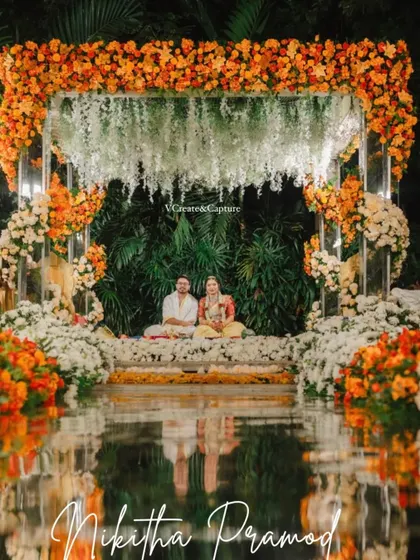 A beautiful Muhurtham mandap where a cascade of white florals meets a vibrant border of orange marigolds, all reflected in the water below.