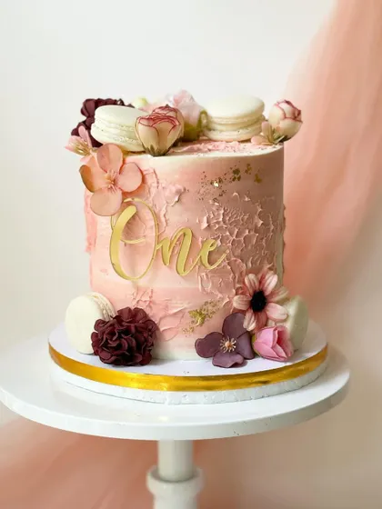 A gorgeous first birthday cake with a textured pink buttercream finish. It's decorated with macarons, delicate silk flowers, and a gold "One" topper.