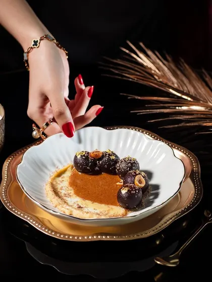 An elegant dessert shot of Kala Gulab Jamun for Club Jolies. The composition is luxurious, with gold-rimmed plates and soft candlelight, perfectly matching the fine dining and fusion concept of the dish.