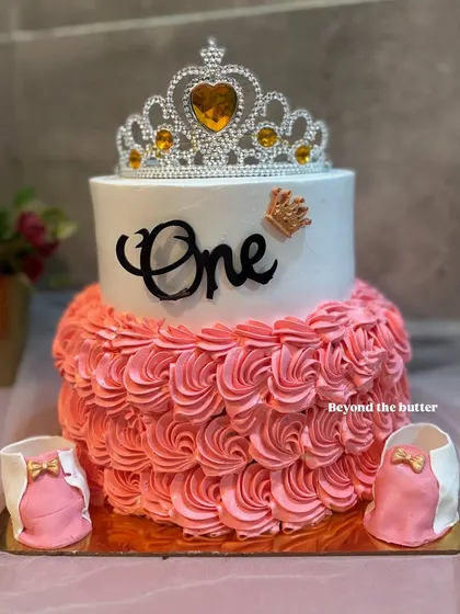 A first birthday cake fit for a little princess. This two tier creation features a sparkling tiara, delicate pink buttercream ruffles, and tiny edible baby booties.