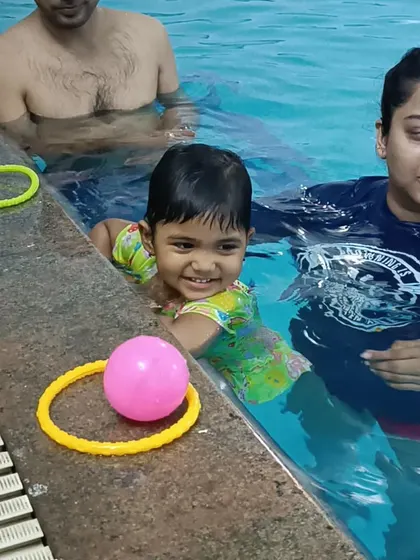A smiling toddler plays with toys by the pool edge during an infant swim class, showing the fun and safe environment.