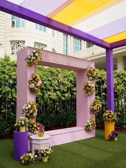 A 'Sun-kissed memories' photo frame for an outdoor Haldi or Mehendi. The simple pink frame is adorned with small floral bouquets, creating a perfect spot for guests to capture joyful moments in the sun.
