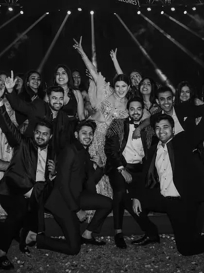 A black and white group photo of the wedding party on the Sangeet stage, full of energy and fun poses.