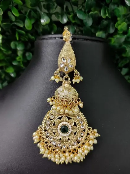 A grand, oversized maang tikka with a passa-jhoomar style. The intricate design features a central green stone and is adorned with numerous pearls.