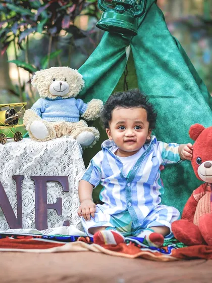 A one-year-old boy sits proudly next to his birthday setup, complete with a "ONE" sign, teddy bears, and a toy plane, ready for his adventure to begin.