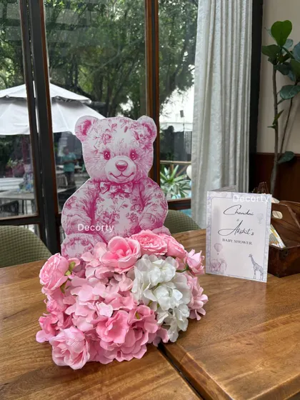 A charming table centerpiece for a Dior-themed baby shower, featuring a pink floral arrangement and a custom-printed teddy bear prop.