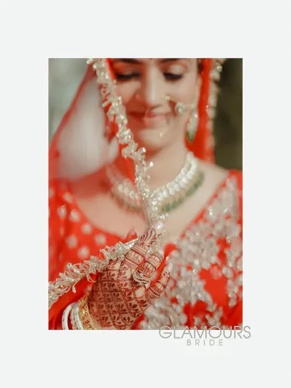 A close-up, artistic shot of the bride peeking through her ghunghat (veil). The focus is on her henna-adorned hand and her gentle smile.