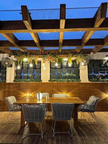 A view of a terrace dining setup under a wooden pergola with a glass roof. The space is enhanced by vertical wall planters and hanging greenery, creating a perfect setting for an evening meal.