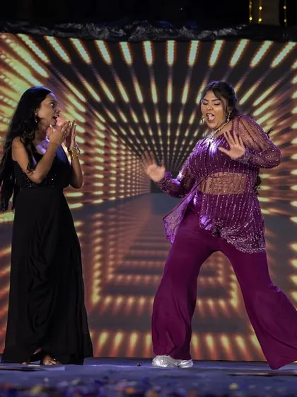 Two women share a fun, energetic dance performance on the Sangeet stage, with a dynamic light show in the background.