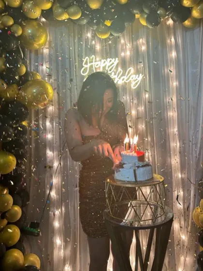 The moment the candles are lit is always magical. This photo captures the birthday girl cutting her cake in front of a sparkling shimmer wall and neon sign, surrounded by a cascade of black and gold balloons.