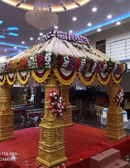 A classic four-pillared wedding mandap decorated with vibrant yellow and red flower garlands, set under a beautiful chandelier in a convention hall.