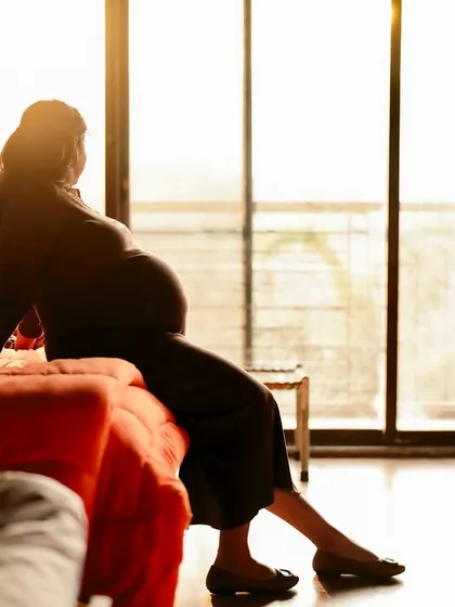 A beautiful silhouette of the mom-to-be sitting on a bed, looking out a large window at sunrise or sunset. It’s a peaceful and contemplative indoor portrait.
