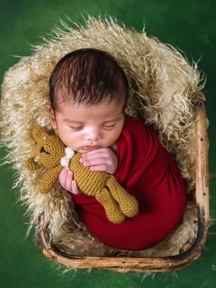 A sweet newborn in a red swaddle, cuddling a small knitted teddy bear while sleeping in a rustic basket.