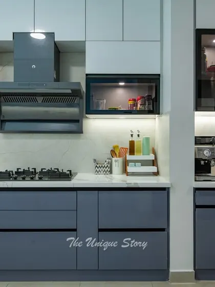 The cooking zone features a built-in hob and chimney, with small glass-shuttered cabinets on either side for spices and essentials. The under-cabinet lighting ensures a well-lit workspace.