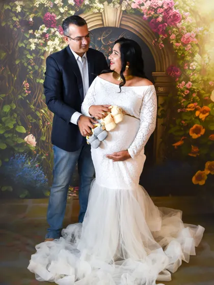 A sweet studio portrait of a couple expecting twins. They are holding two pairs of baby shoes, sharing a happy moment in front of our floral arch backdrop.