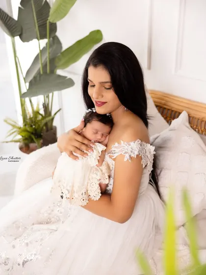 A mother in an elegant white gown, holding her sleeping newborn. The natural light and indoor plants create a serene and peaceful setting.