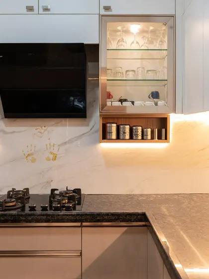 A close-up of a kitchen corner, showing a glass-front cabinet for displaying glassware and a well-lit open shelf for frequently used items. The handprints on the marble backsplash add a sweet, personal touch.