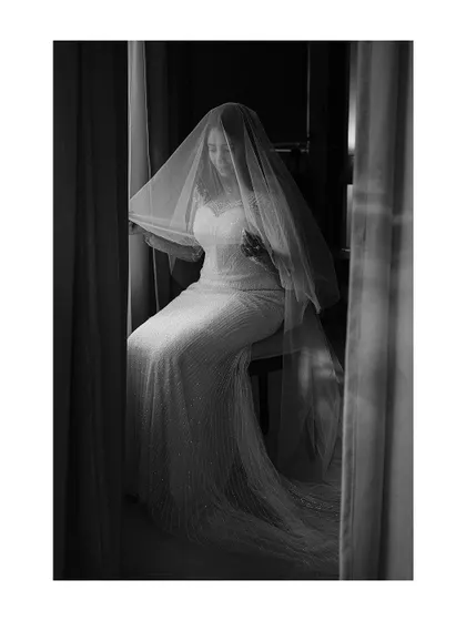 A moody black and white portrait of a bride seated by a window, her face obscured by her veil. The image is full of atmosphere and quiet emotion.