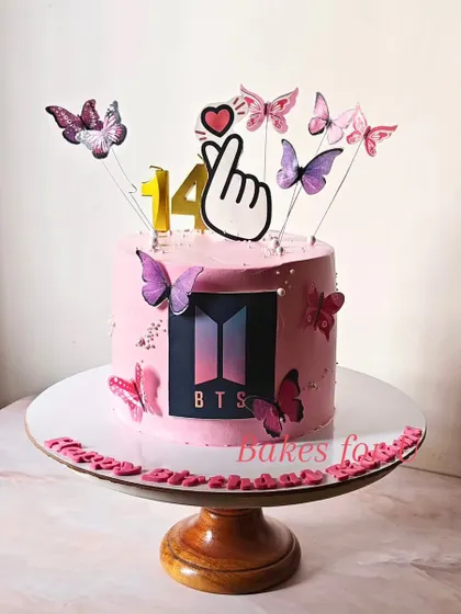 A BTS Army cake for a 14th birthday, decorated in the band's signature pink and black colors. The design includes the BTS logo, butterflies, and the iconic finger heart gesture.