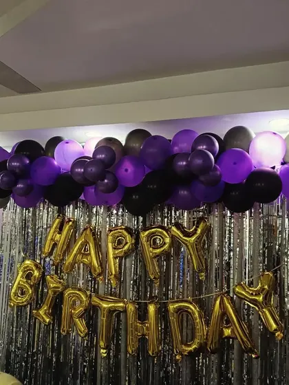 A purple and black balloon garland over a silver tinsel curtain with gold 'Happy Birthday' foil letters.
