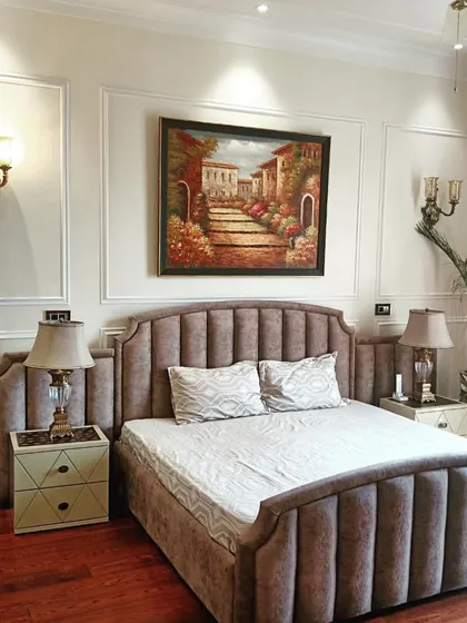 An elegant bedroom with a classic aesthetic, featuring a custom-made bed with a curved and fluted beige headboard. The room is furnished with matching bedside tables and traditional wall sconces, creating a warm and inviting atmosphere.