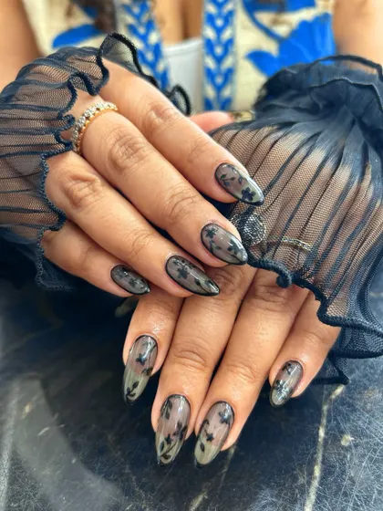 An example of elegant and sophisticated nail art. These almond-shaped nails feature a sheer, translucent base with delicate black floral patterns, creating a beautiful lace-like effect that is both subtle and intricate.