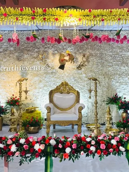 A classic stage for a traditional function, featuring a backdrop made of white flowers. The setup is framed with garlands of jasmine and roses, with a single ornate chair at the center.