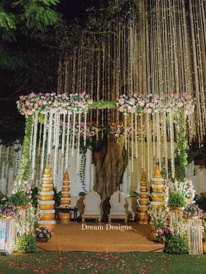 A full view of a traditional mandap setup under a canopy of twinkling lights. The design incorporates classic golden pillars and a profusion of pink and white flowers, creating a sacred and opulent space for the wedding ceremony.