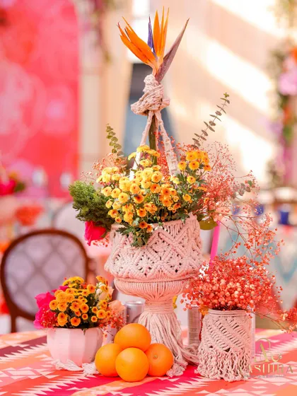 A whimsical centerpiece featuring a macrame plant hanger holding a pot of marigolds, surrounded by fresh oranges and other floral arrangements.