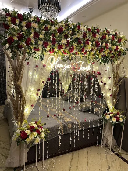 A classic wedding night decoration with a bed canopy made of red and white flowers, pampas grass, and a custom neon sign.