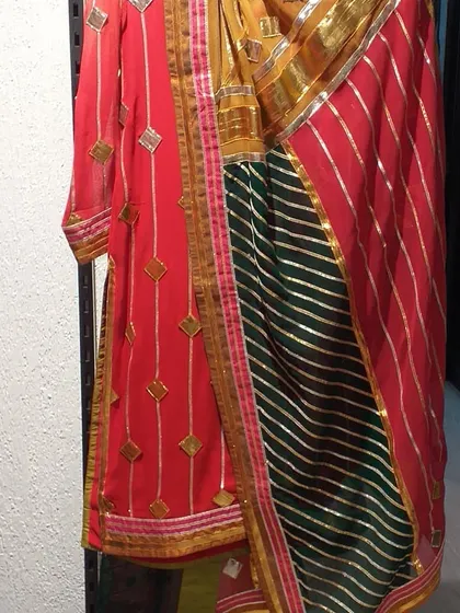 A vibrant Gota Patti suit by designer Preeti S Kapoor, featuring a red kurta and a multi-colored dupatta with green and red stripes. This is a perfect example of the renowned designer labels available in our outlet.