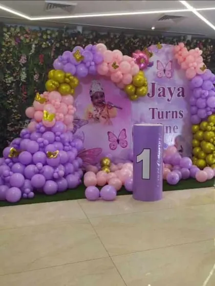 A butterfly and Krishna-themed first birthday party. This decoration includes a large balloon arrangement in purple and pink, a custom backdrop, and a light-up number "1".