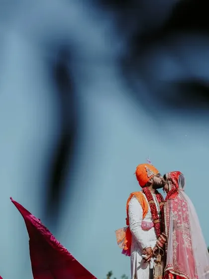 An artistic and creative angle of the Varmala ceremony, capturing a kiss from a low angle against a clear blue sky.
