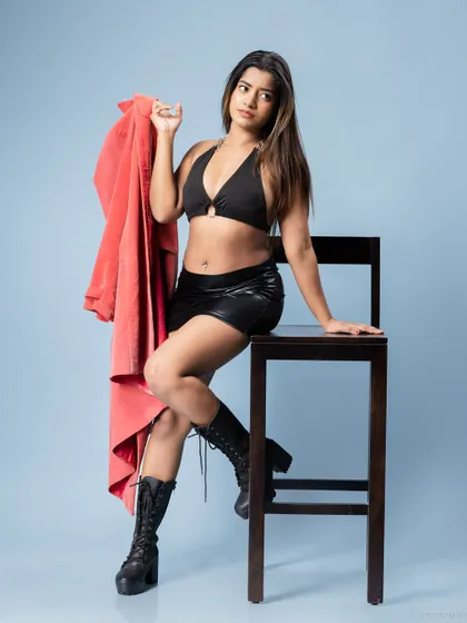 A model seated on a high stool, wearing a black bikini top, leather-look mini skirt, and combat boots, holding a red jacket. This shot combines different textures and colors for a modern, edgy portfolio look.