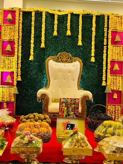 A full view of a Seemantham stage, featuring a colorful backdrop with hanging marigold garlands and brass bells. The foreground is filled with a wide variety of traditional sweets and offerings.