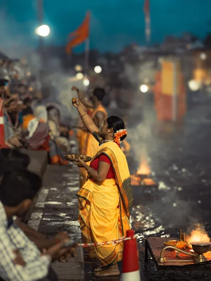 A woman in a yellow saree offers prayers at the Ganga ghat. The smoke from the incense and the diyas creates a spiritual and serene atmosphere.