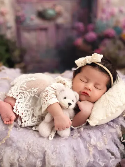A sleeping angel. This newborn girl is dressed in a lace romper, cuddling a tiny teddy bear in a magical, lavender-themed fairy setup.
