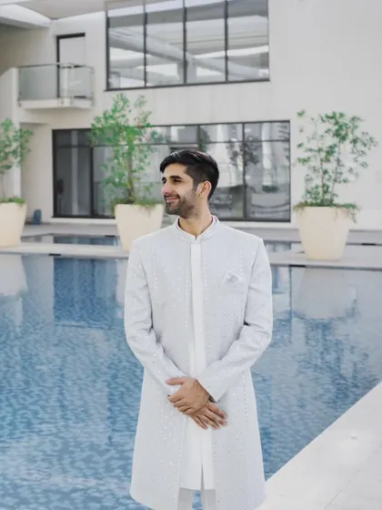 A portrait of the groom looking dapper in his light-colored sherwani, standing by the poolside of a modern hotel venue before his ring ceremony.