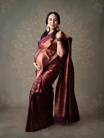 A graceful solo portrait. The mom-to-be's pose highlights the rich texture and beautiful drape of her maroon silk saree against a mandala-patterned backdrop.