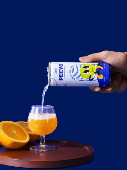 An action shot showing the citrus soda being poured, creating a sense of refreshment. The bold blue background makes the orange color pop, resulting in a vibrant and energetic beverage photograph.